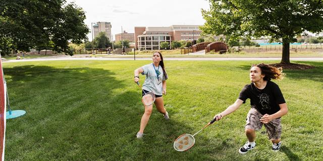 Two people playing badminton on a grassy field with trees and buildings in the background.