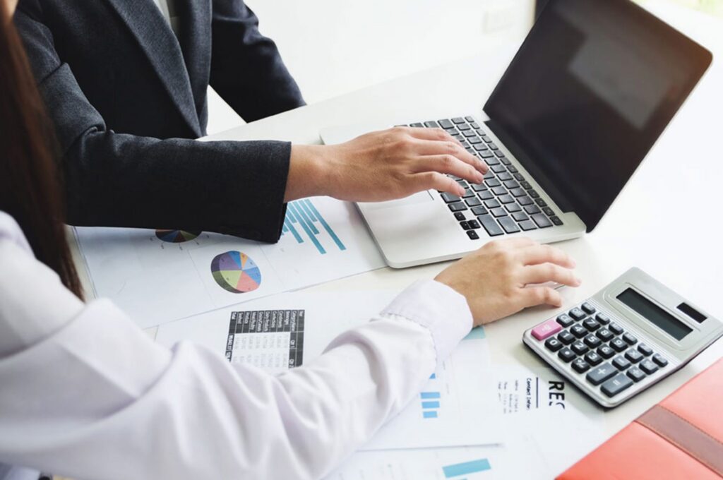 Two people are working together at a table with a laptop, papers with charts, and a calculator.
