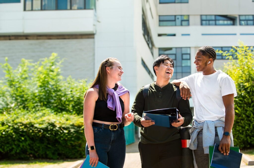 Three people are laughing and talking while holding folders outside a building, surrounded by greenery.