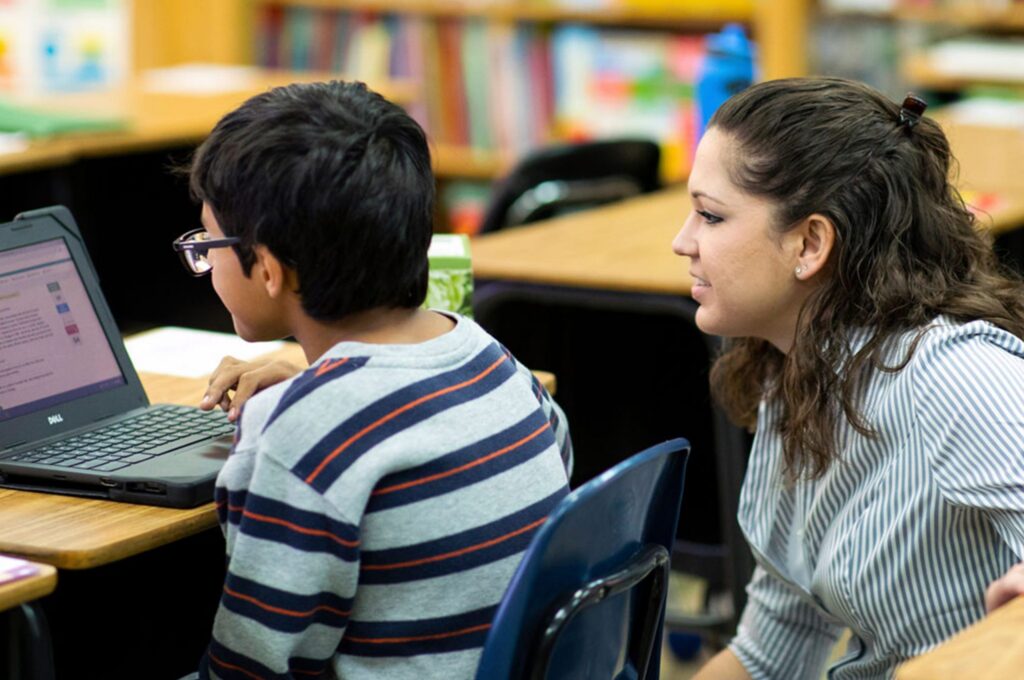 A person assists a child at a laptop in a classroom setting, both focused on the screen.