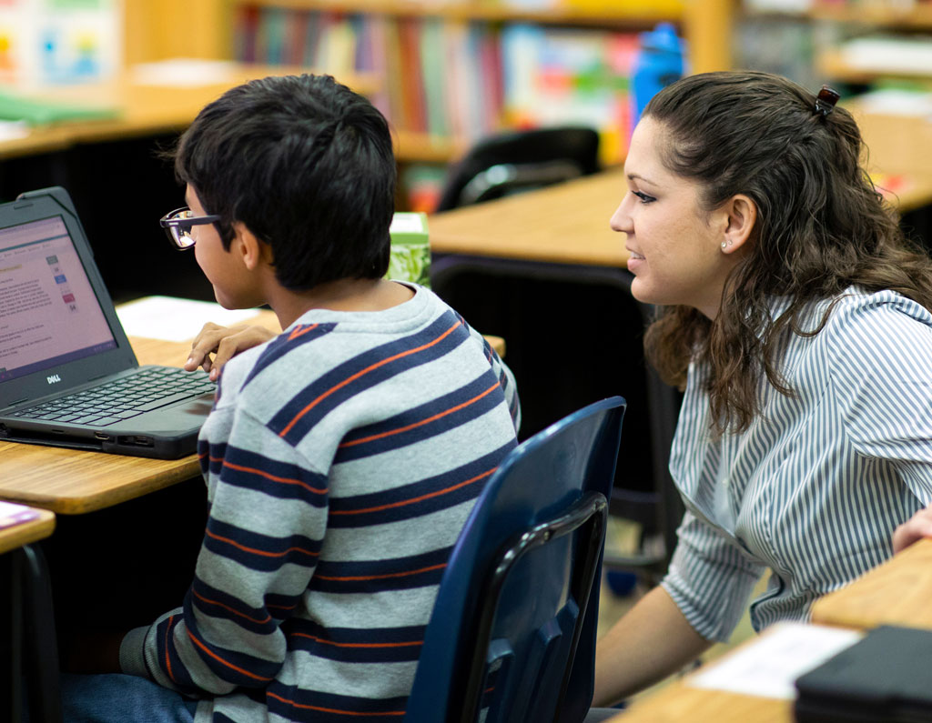 A person assists a student at a laptop in a classroom setting, with bookshelves visible in the background.