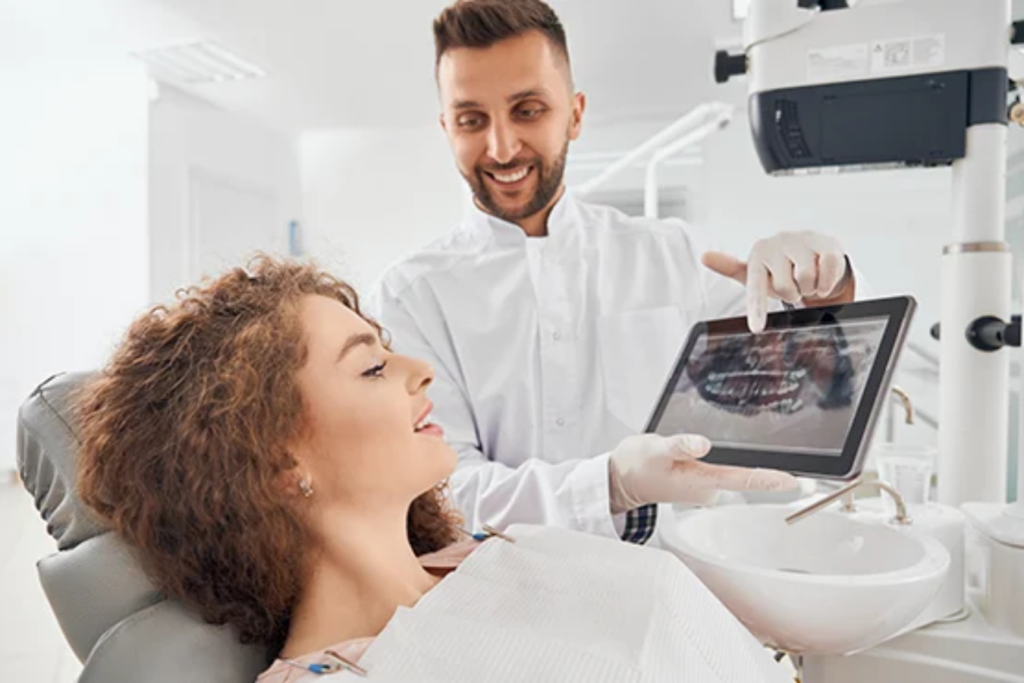 A person in a dental office shows a patient an image on a tablet while they are seated in a dental chair.
