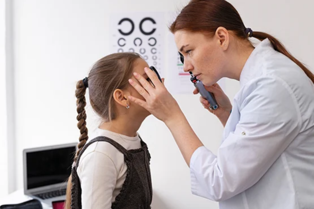 A healthcare professional examines a child's eyes in a medical office, with an eye chart visible in the background.