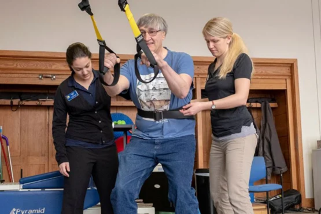 A person uses a harness and straps for support while two others assist them in a rehabilitation setting.