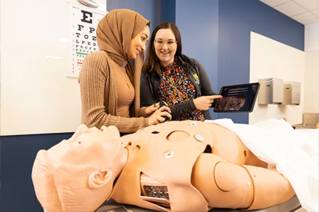 Two people are interacting with a medical training mannequin while one holds a tablet, discussing its features in a classroom setting.