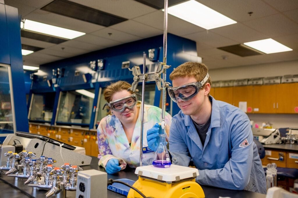 Two people in safety goggles conduct an experiment in a laboratory, focusing on a flask being heated on a scale.