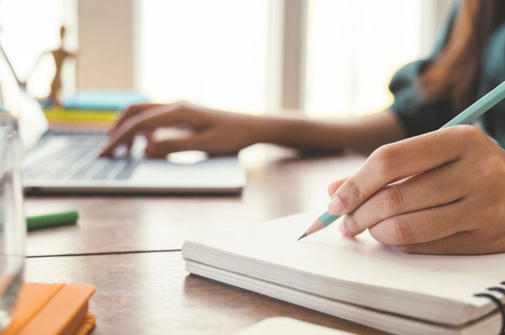 A person is writing in a notebook with a pencil while using a laptop on a wooden desk.