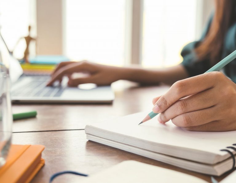 A person writes in a notebook with a pencil while using a laptop on a desk.