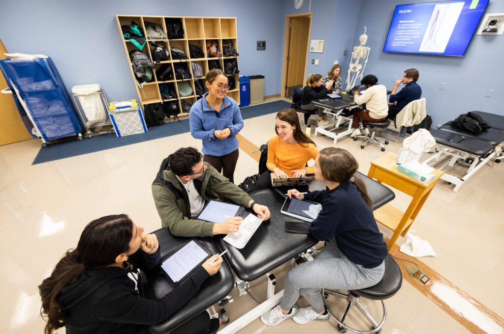 A group of students collaborates at tables in a classroom, with a presentation on anatomy displayed on a screen.