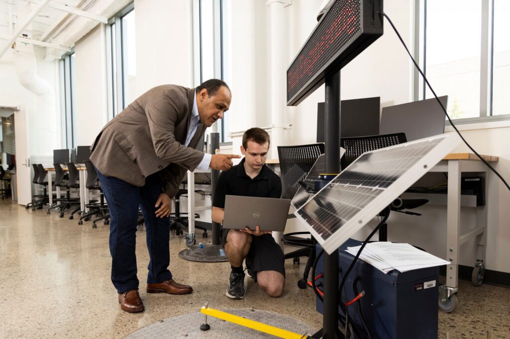 A person points while discussing with another person who is kneeling and using a laptop next to a solar panel setup.