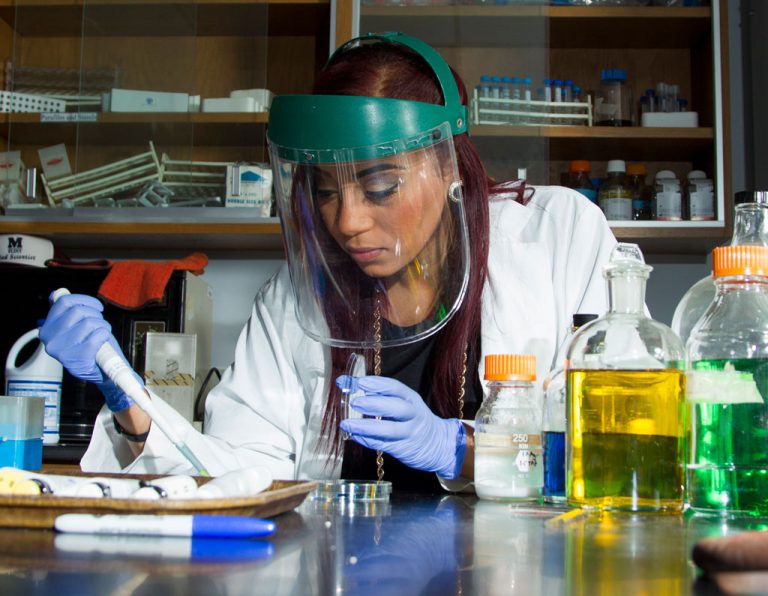 A person in a lab coat and face shield is using a pipette to transfer liquid in a laboratory setting with various colored liquids.