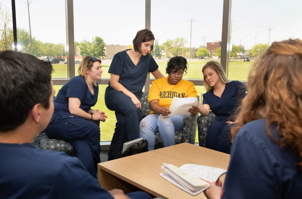 A group of five people in scrubs gather around a table, discussing a document while seated in a well-lit room.