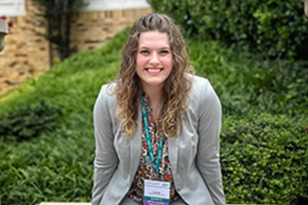 A person smiles while sitting on a bench in front of greenery, wearing a blazer and a name tag.