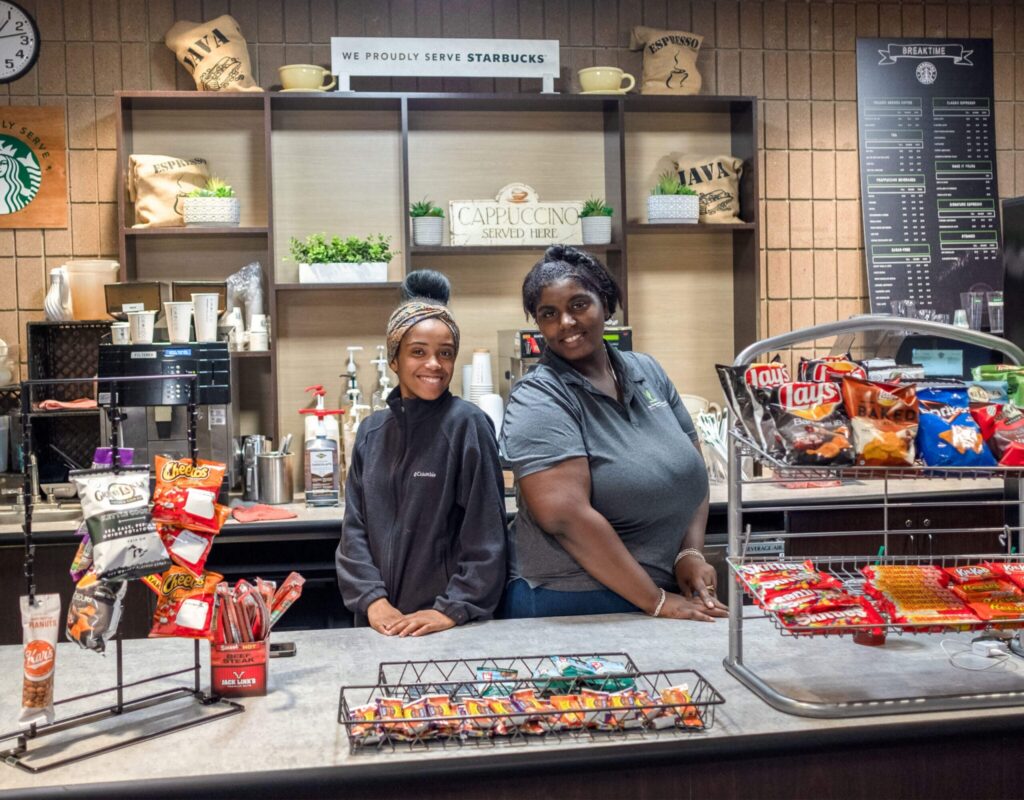 Two people stand behind a counter at a Starbucks, smiling, with snacks and drinks displayed in front of them.