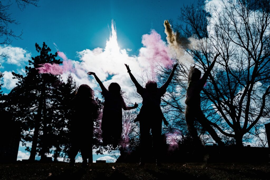 Silhouettes of four people jumping and celebrating outdoors, with colorful powder clouds against a bright sky.