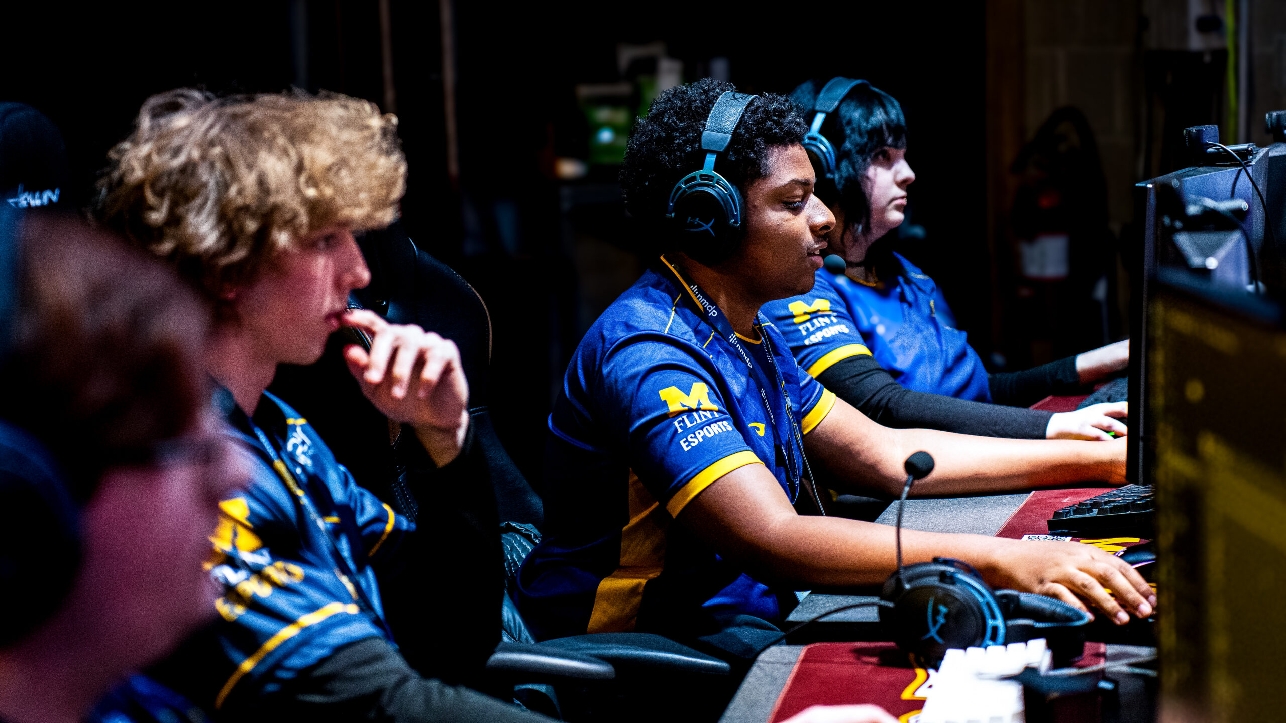 A group of four people wearing headsets and esports jerseys focus on their computer screens during a gaming event.