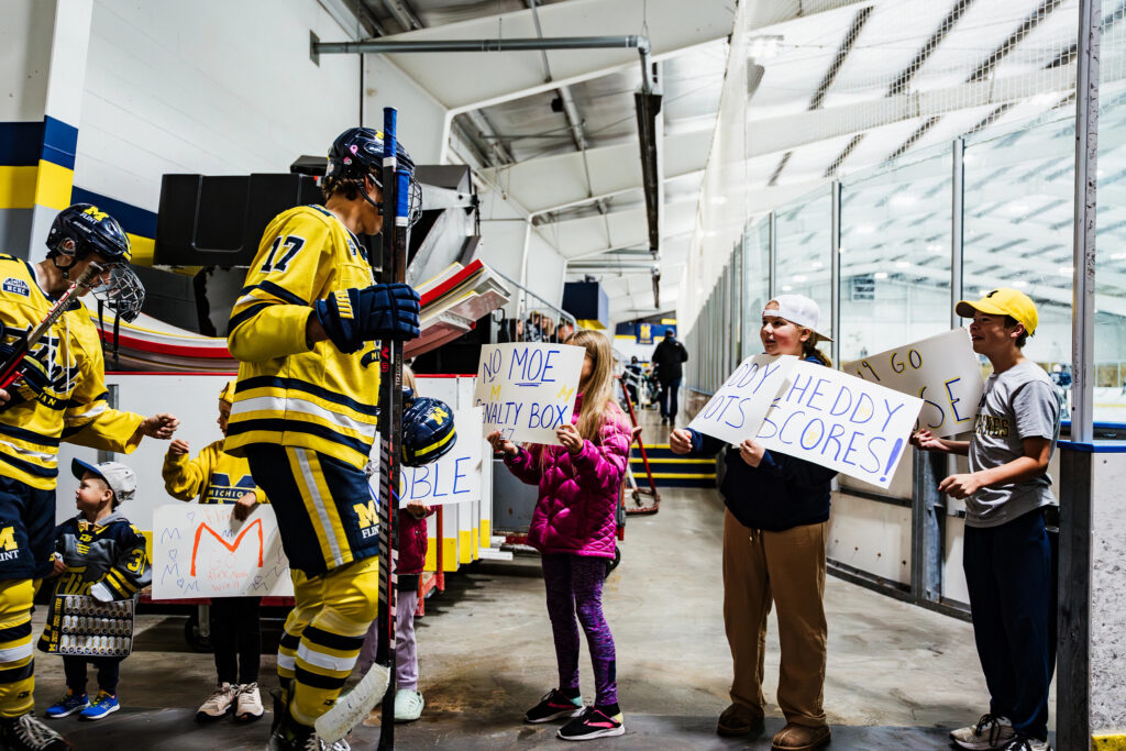 A hockey player in a yellow jersey walks past children holding signs in a rink, cheering and showing support.