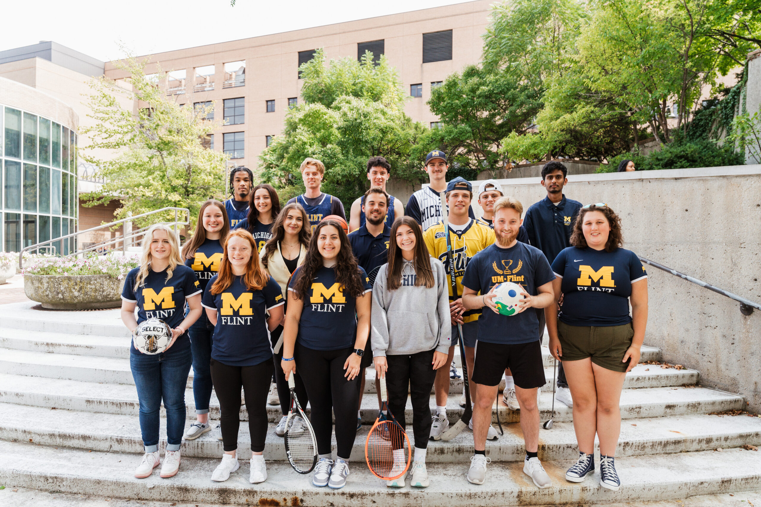 A group of students wearing UM-Flint shirts poses on outdoor steps, holding sports equipment like soccer balls and tennis rackets.