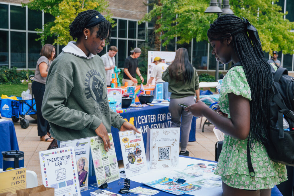 Two people are engaged at a table displaying various informational materials and flyers at an outdoor event.