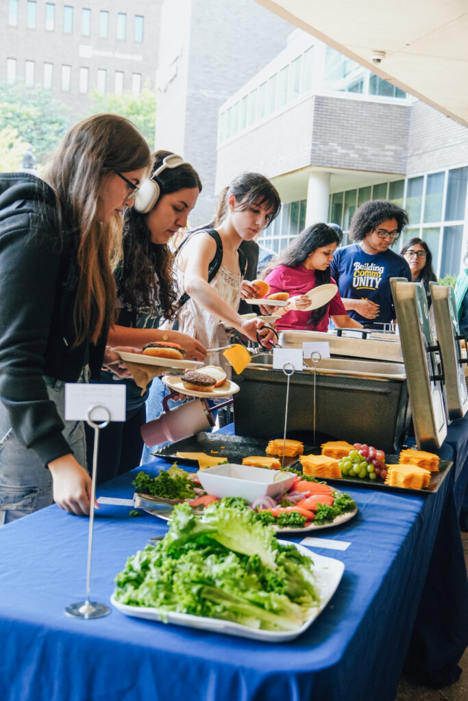 A group of people serving themselves food from a buffet table with various items, including lettuce, cheese, and fruits.