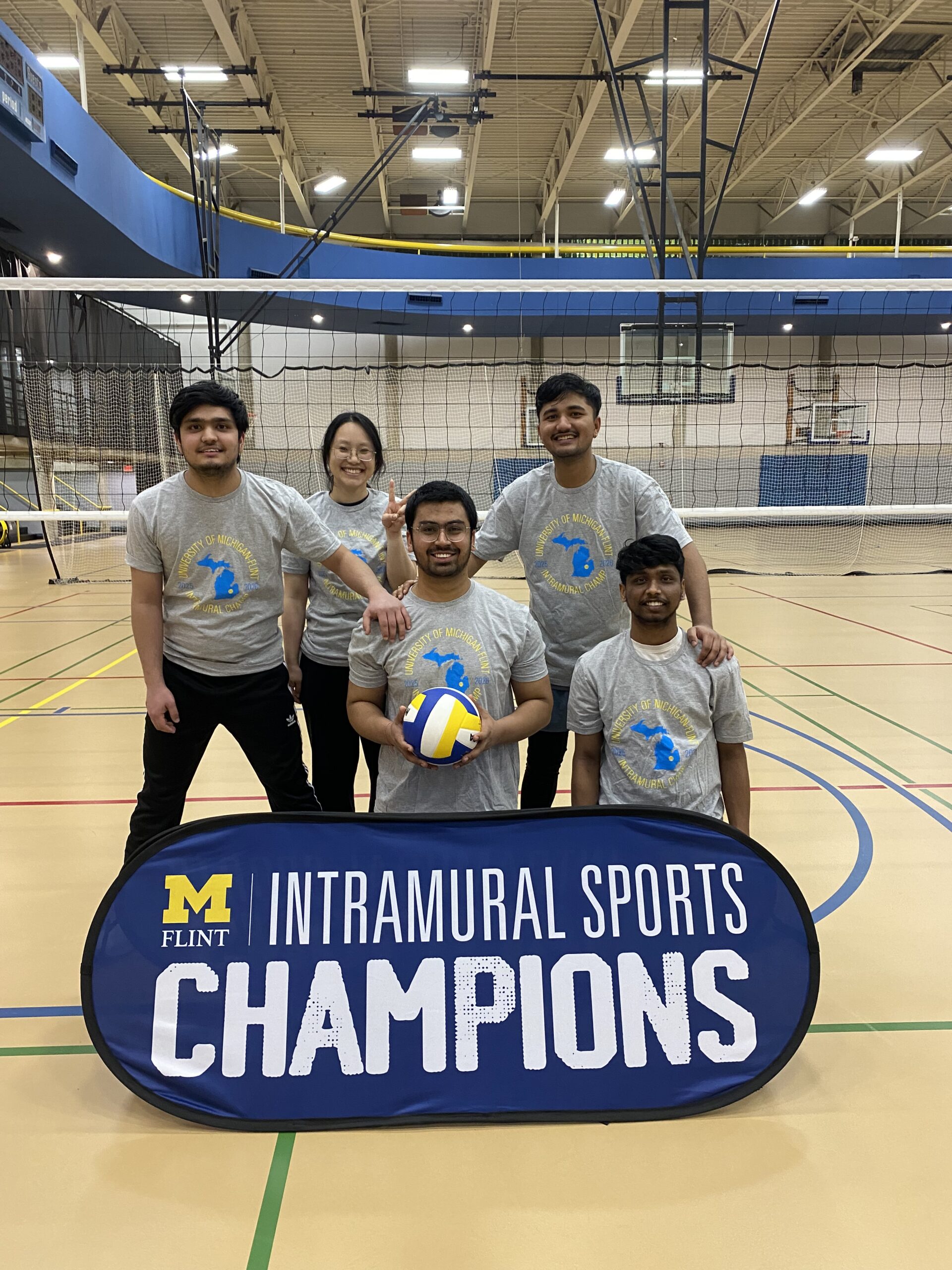 A group of five people poses in a gym with a volleyball and a banner that reads "INTRAMURAL SPORTS CHAMPIONS."
