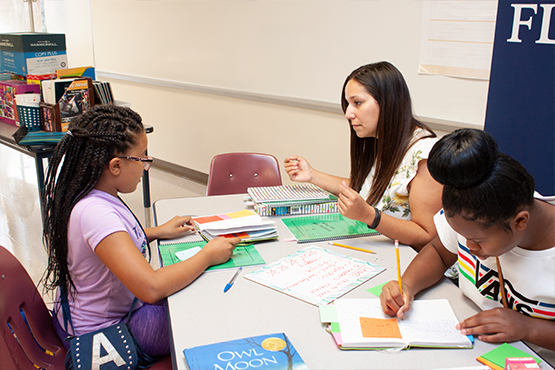 A caregiver helping two children with schoolwork at a table.