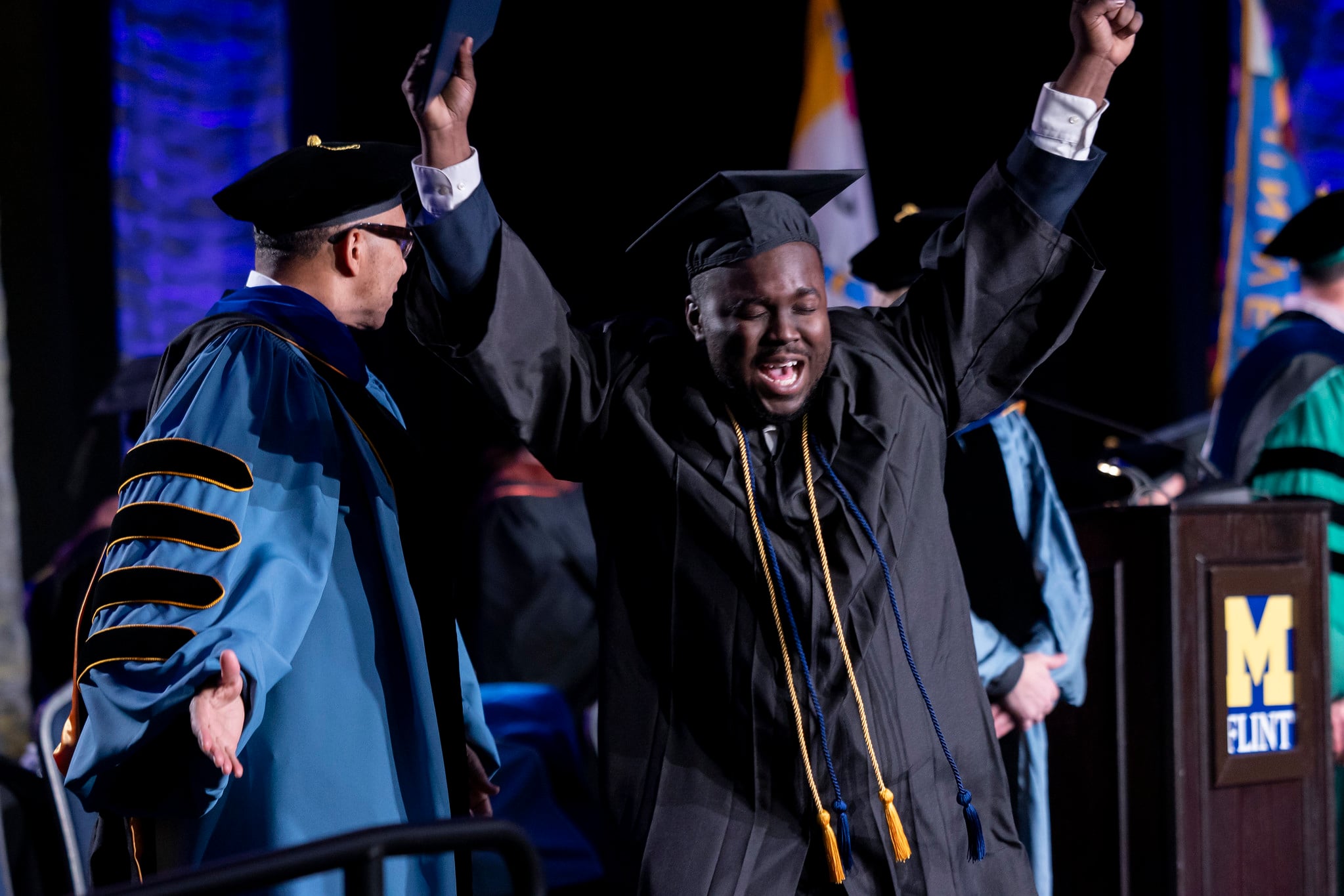 UM-Flint commencement with excited student with arms in the air holding a diploma