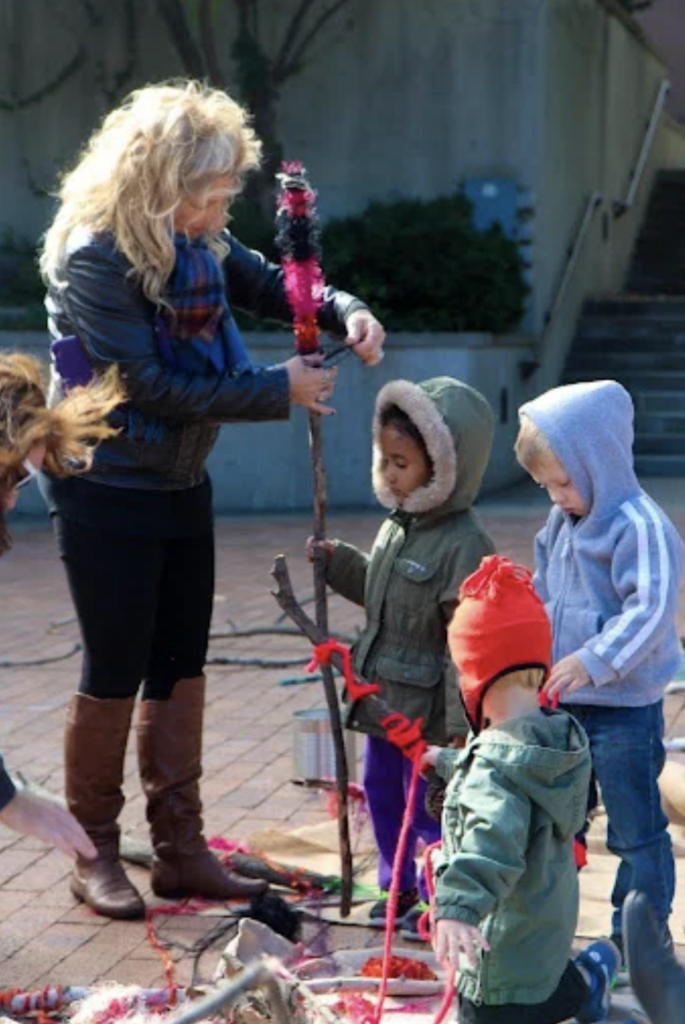 A person assists children in crafting with sticks and colorful materials outdoors.