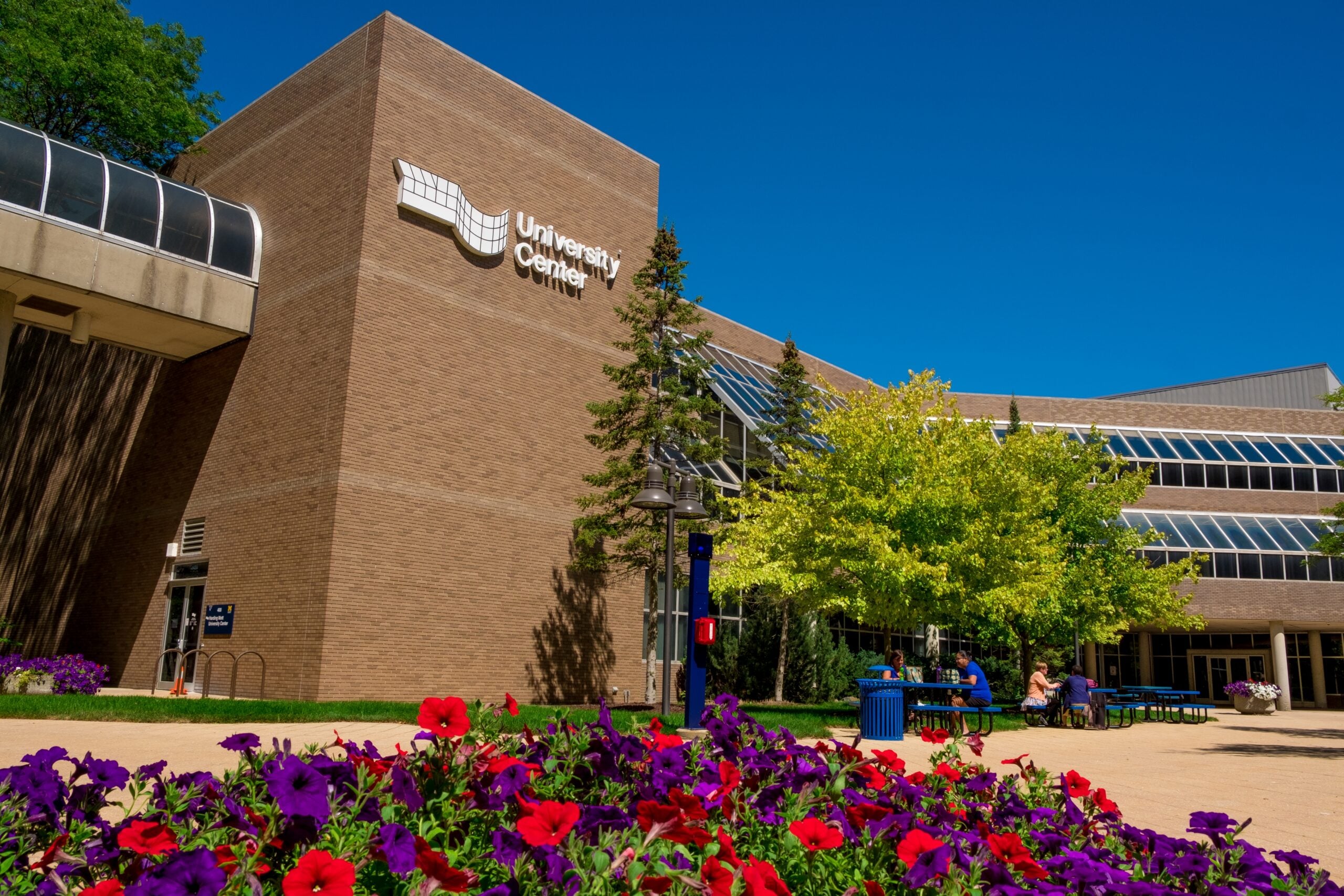 A view of the University Center building with colorful flowers in the foreground and people sitting at tables nearby.