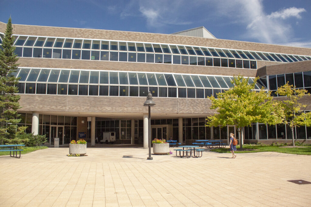 A person walks past a modern building with large glass windows and outdoor seating areas surrounded by trees and flowers.