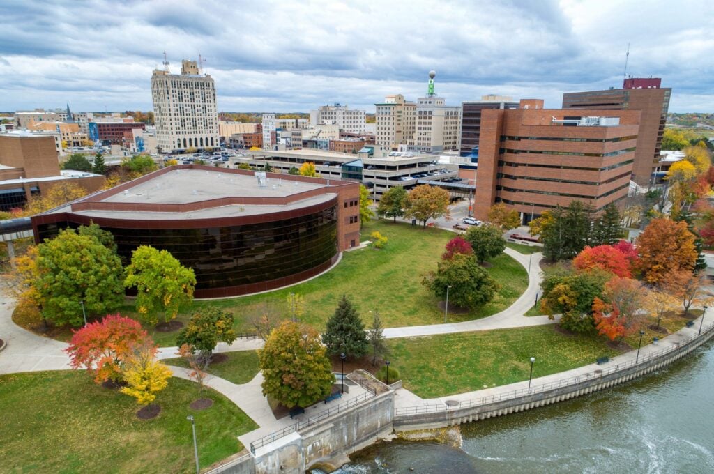 Aerial view of a cityscape featuring a park with colorful autumn trees, surrounded by buildings and a river.