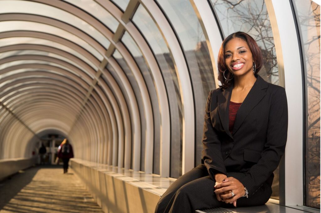A person in a suit smiles while sitting on a ledge in a modern, curved glass walkway, with another person walking in the background.