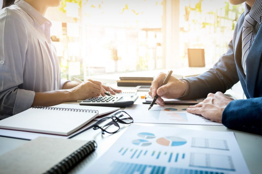 Two people are working at a desk, one using a calculator and the other writing on a document with charts and graphs.
