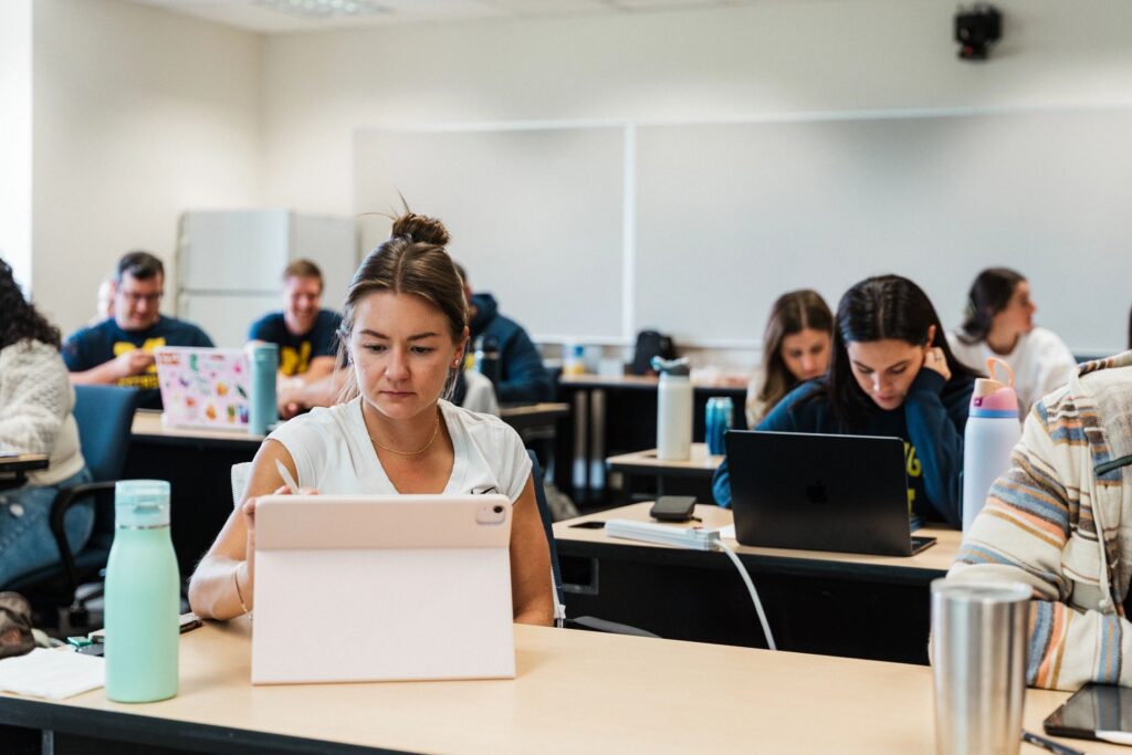 A person sits at a desk using a tablet in a classroom filled with students working on laptops and other devices.