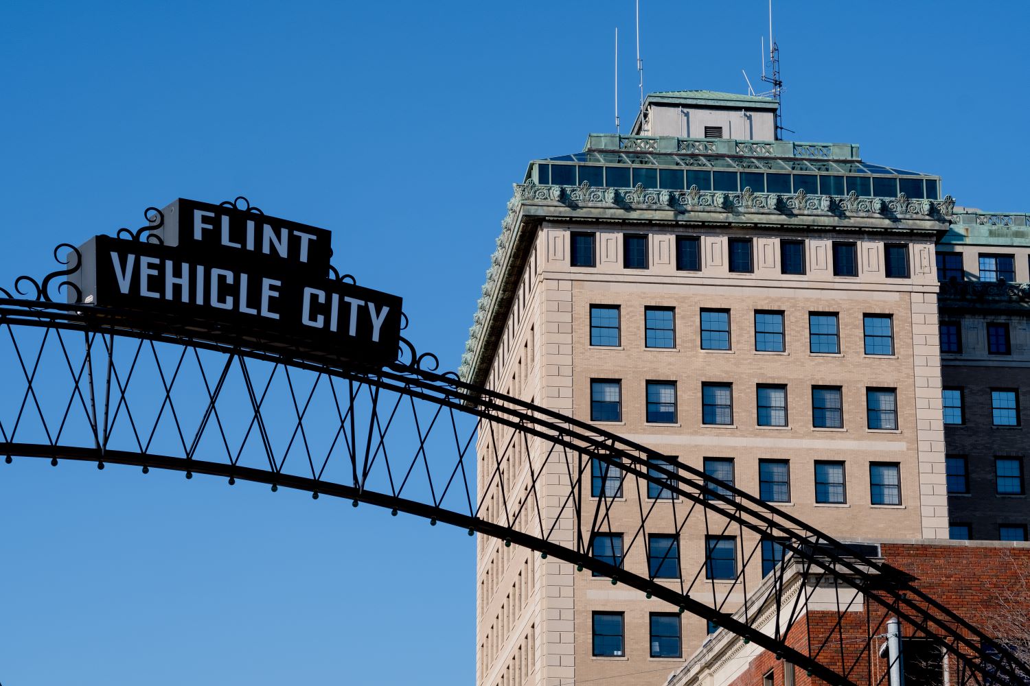 Sign reading "Flint Vehicle City" on an archway in front of a historic building against a clear blue sky.