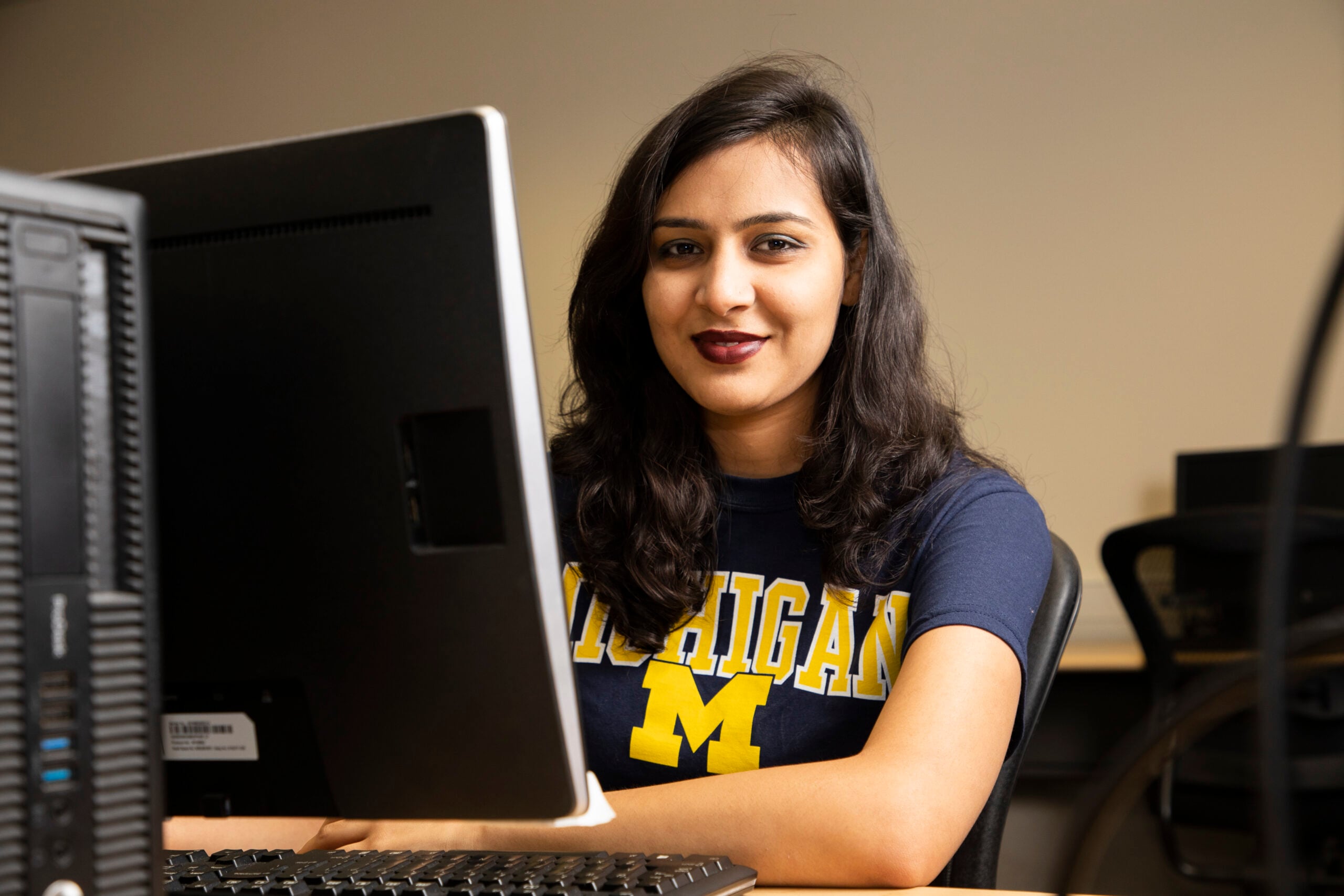 A person sits at a computer, smiling while wearing a shirt with "Michigan" printed on it.