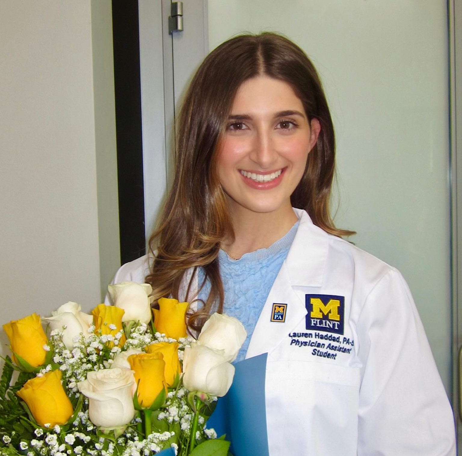 Lauren Haddad in a white lab coat holds a bouquet of yellow and white roses and smiles, standing in front of a light background.