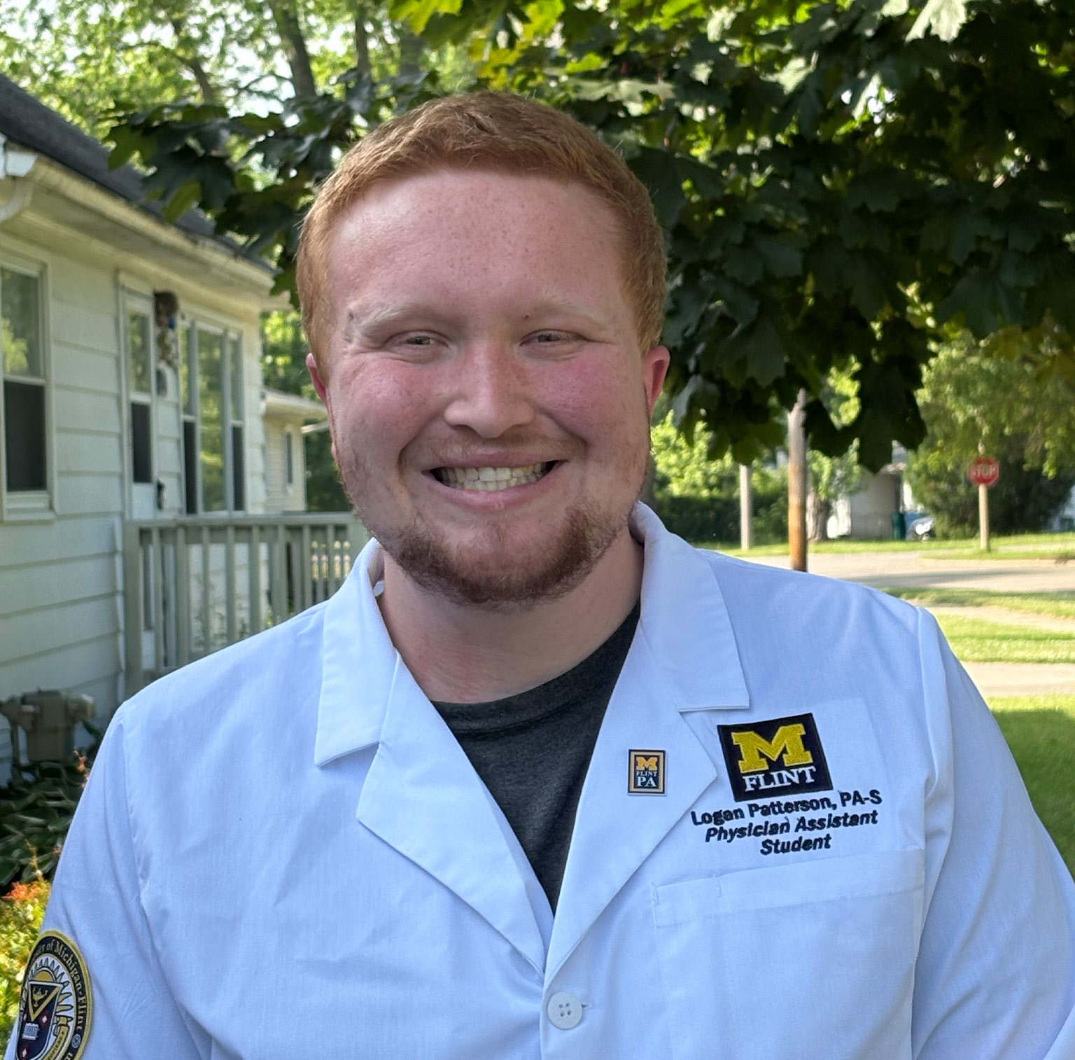 Logan Patterson in a white lab coat smiles outdoors, with a house and trees in the background.