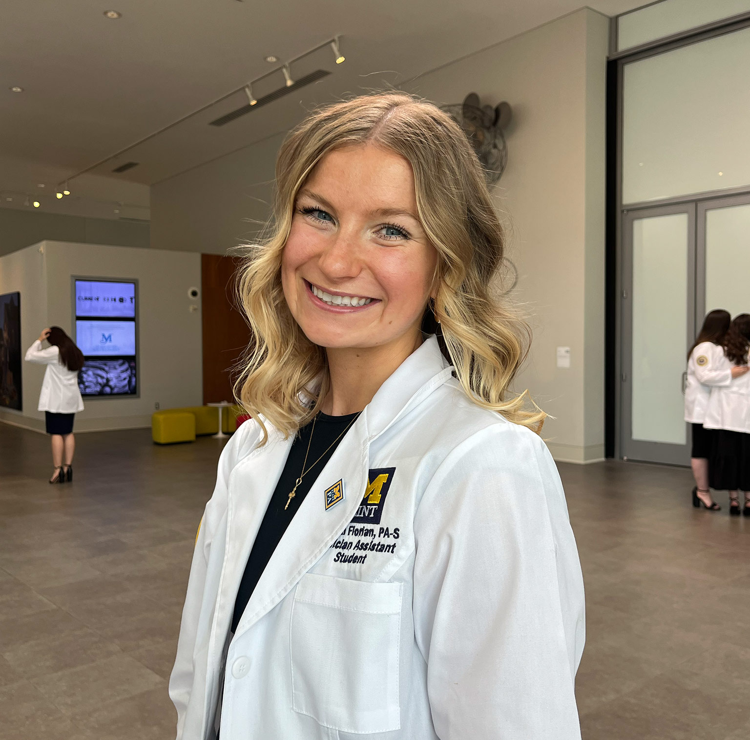 Meghan Florian in a white lab coat smiles in a modern indoor setting, with others in lab coats visible in the background.