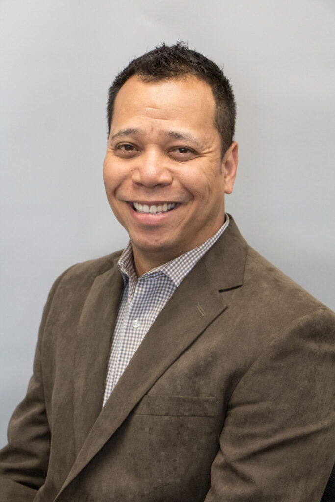 Michael Sartor smiling while wearing a brown blazer over a checkered shirt, set against a gray background.