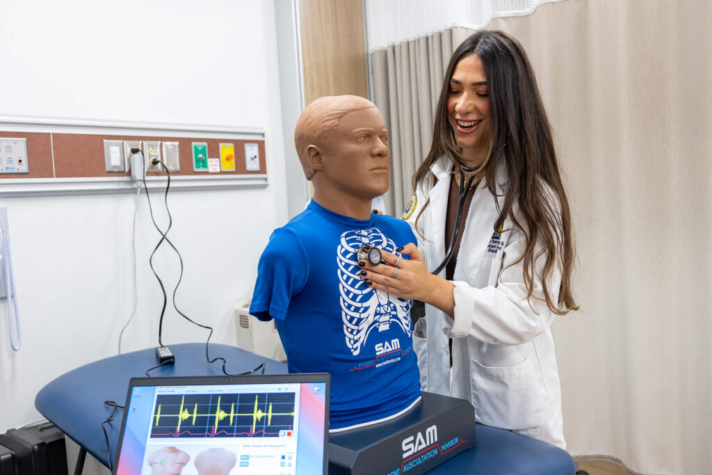 A person in a lab coat uses a stethoscope on a medical training mannequin with a blue shirt displaying a skeleton design.