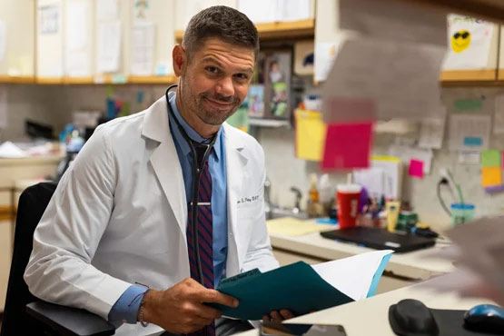 A person in a white lab coat smiles while holding a folder at a desk cluttered with papers and office supplies.