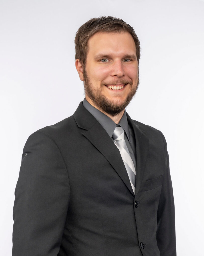 Reed Muehler in a black suit and gray tie smiles against a plain white background.