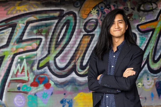 Mitch Sansiribhan stands confidently with arms crossed in front of a colorful graffiti wall that says "Flint."