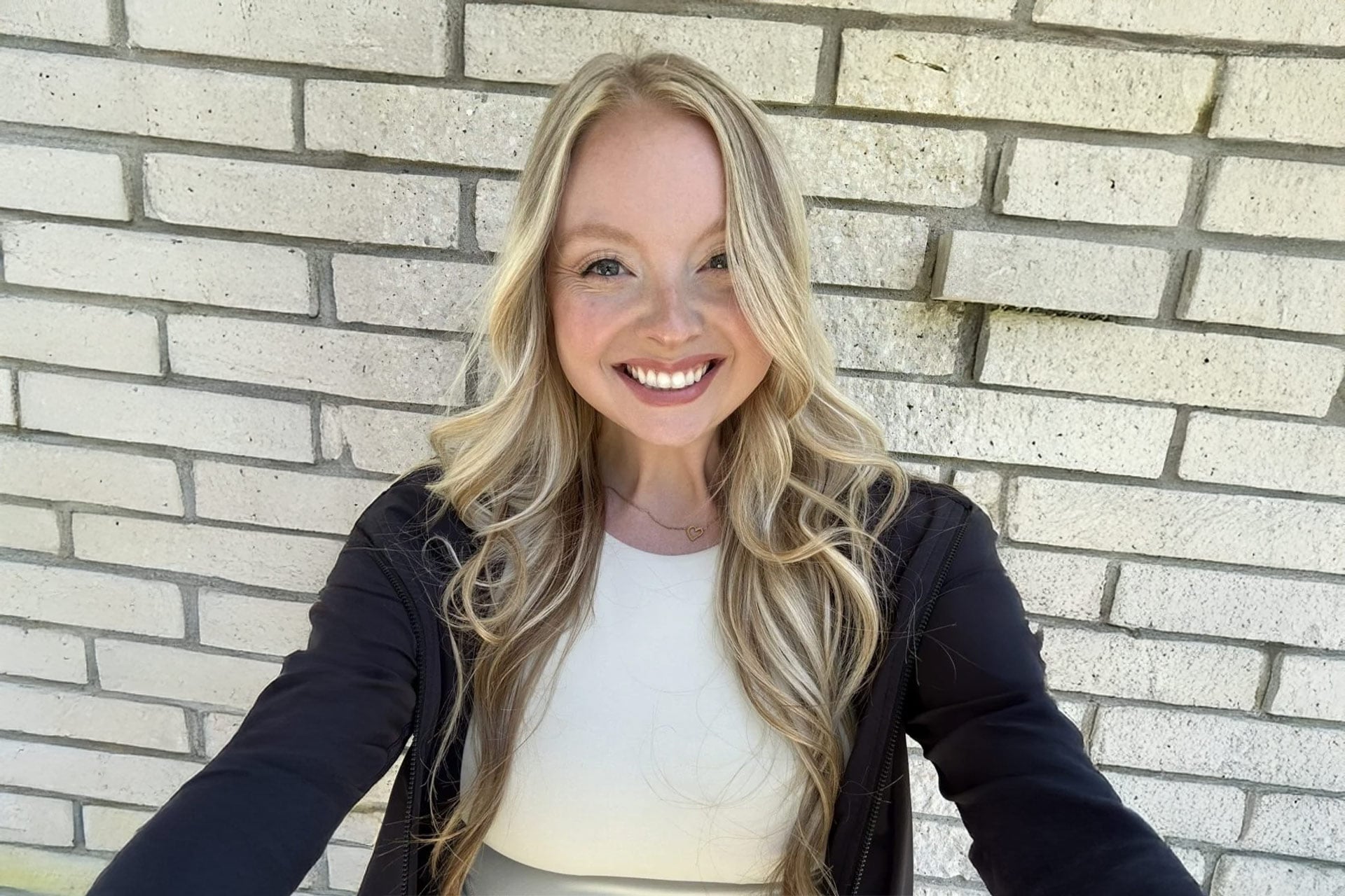 Taylor Maples smiles while taking a selfie in front of a light-colored brick wall.