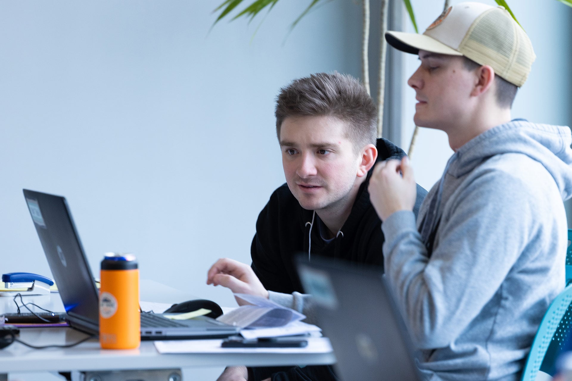 Two people are engaged in discussion while working on a laptop at a table, with a water bottle and papers nearby.