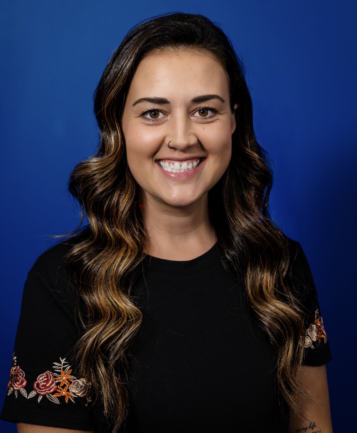 Caela W. with long, wavy hair smiles against a blue background.