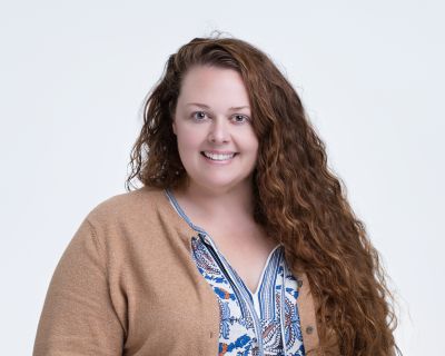Jennifer C. with long, curly hair smiles while wearing a patterned top and a cardigan against a plain background.