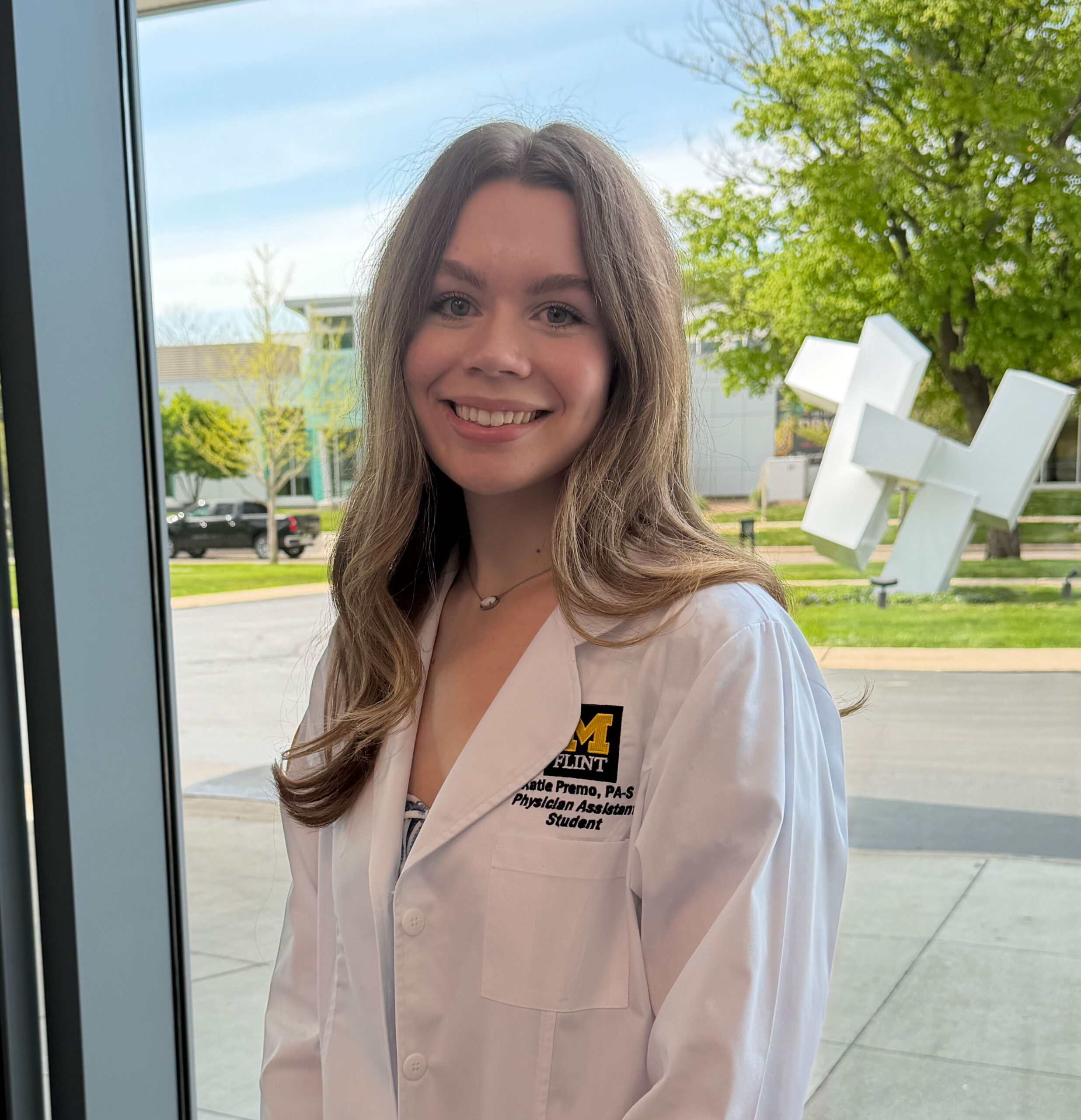 Katie P. in a white lab coat smiles while standing near a window, with greenery and a sculpture visible outside.