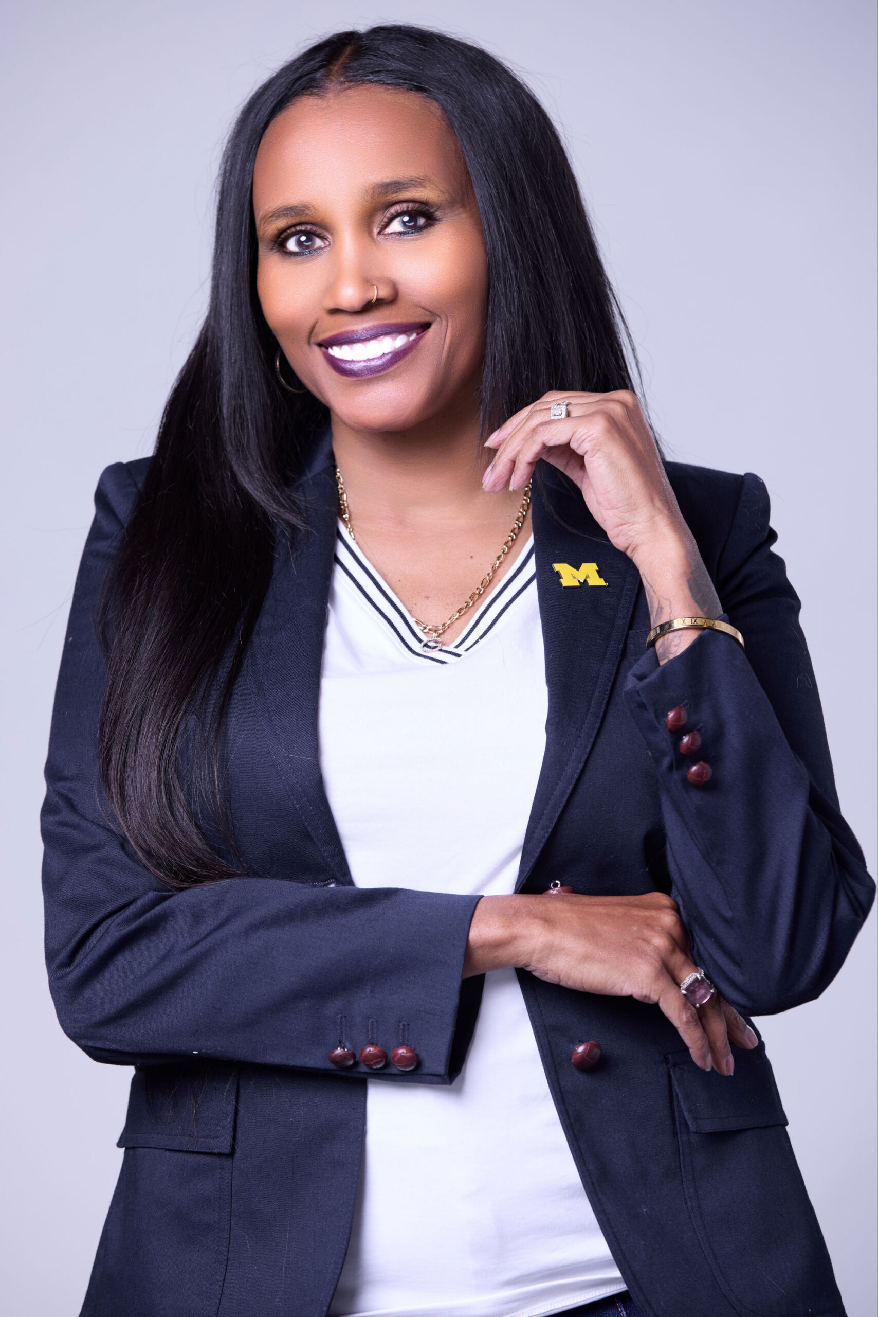 Seana G. in a blazer with a university logo smiles and poses with one hand on their chin and the other crossed over their arm.